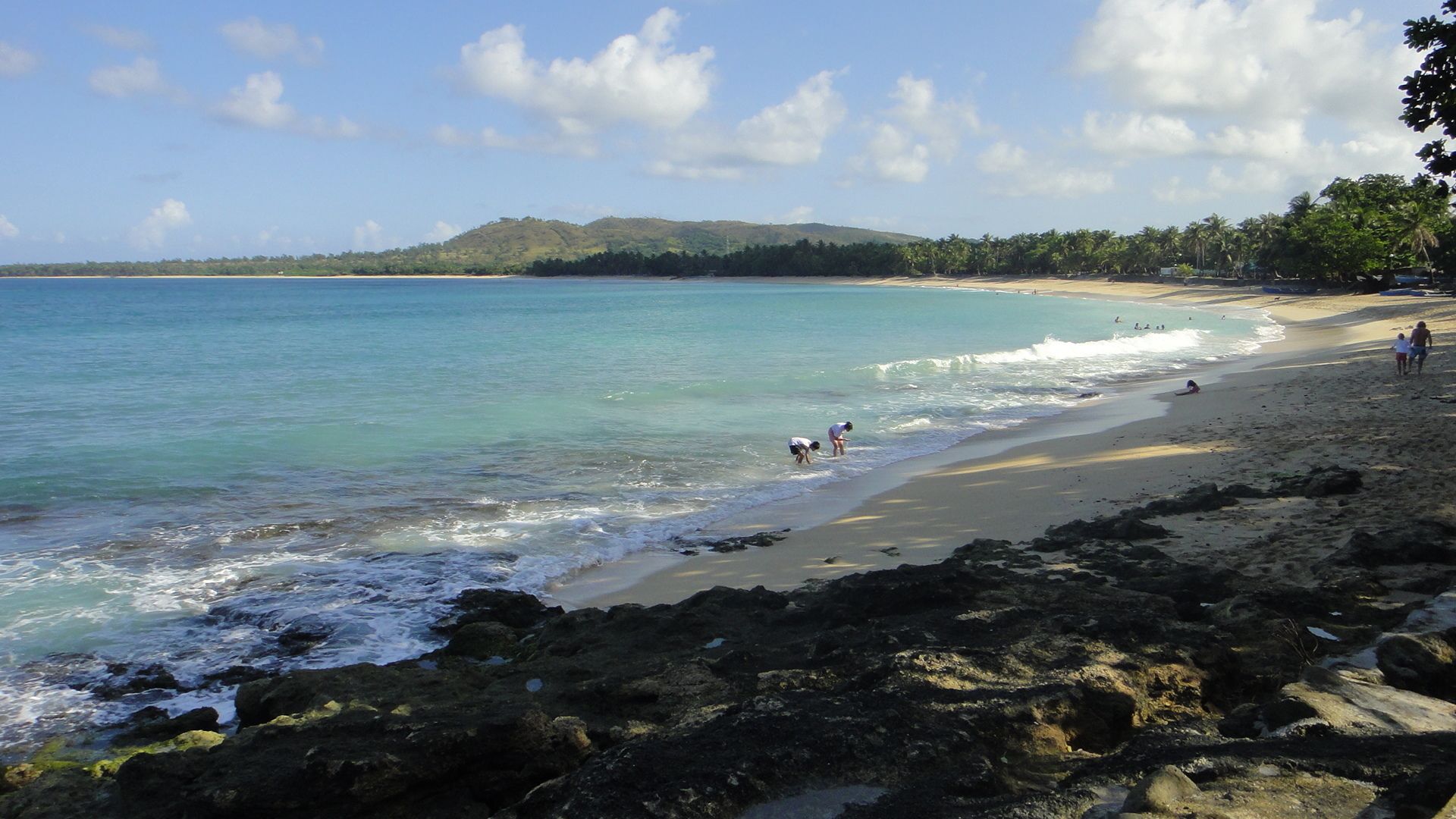 Une plage bordée d’une eau turquoise et limpide, d’une côte rocheuse et d’une colline qui se dessine à l’horizon.