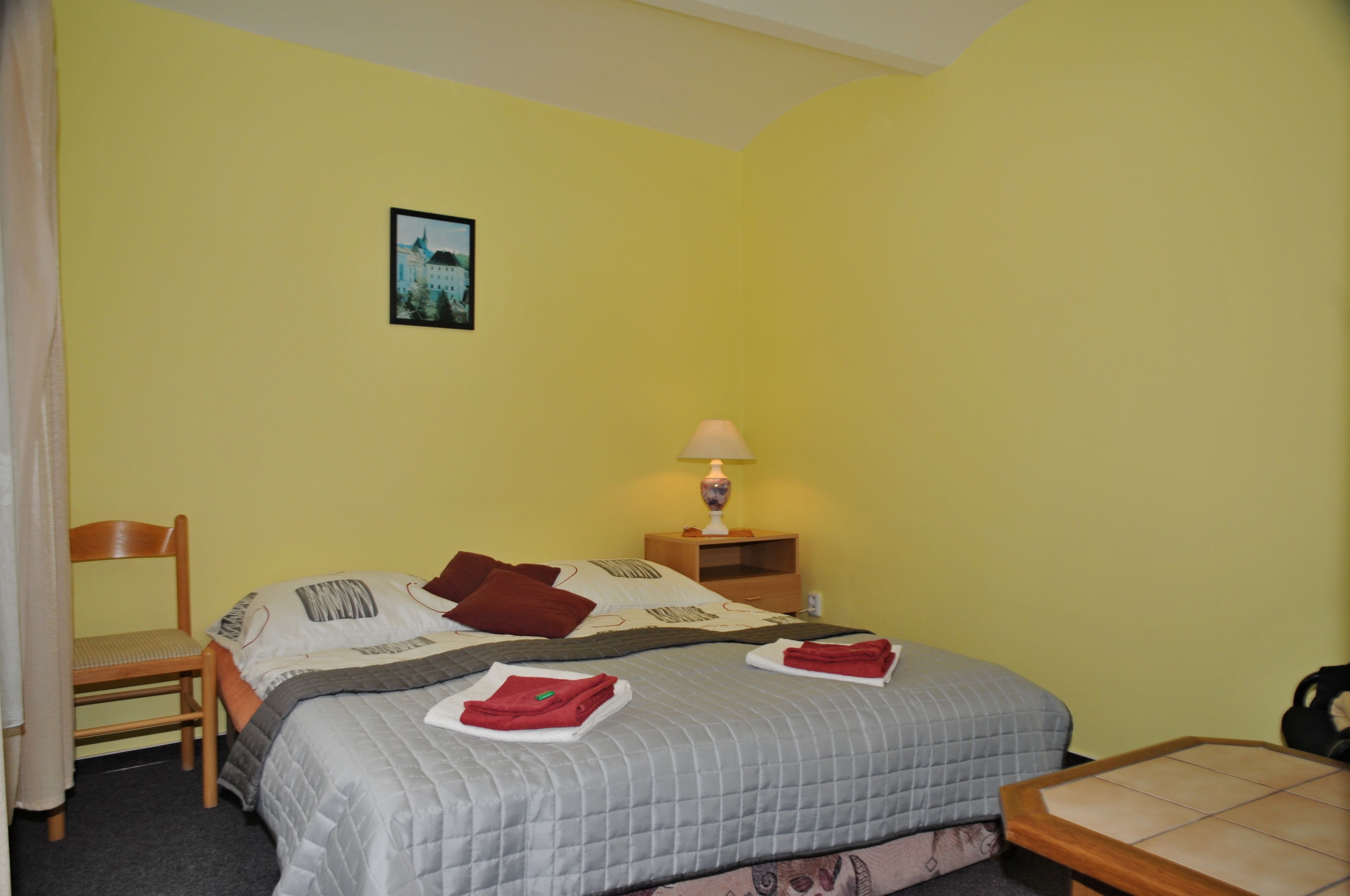 Habitación de hotel con cama, una silla, mesita de noche con lámpara y un cuadro en la pared.
