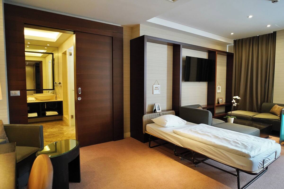 A hotel room with a bed, a sofa, a small table, a TV, and a bathroom with a mirror and sink.