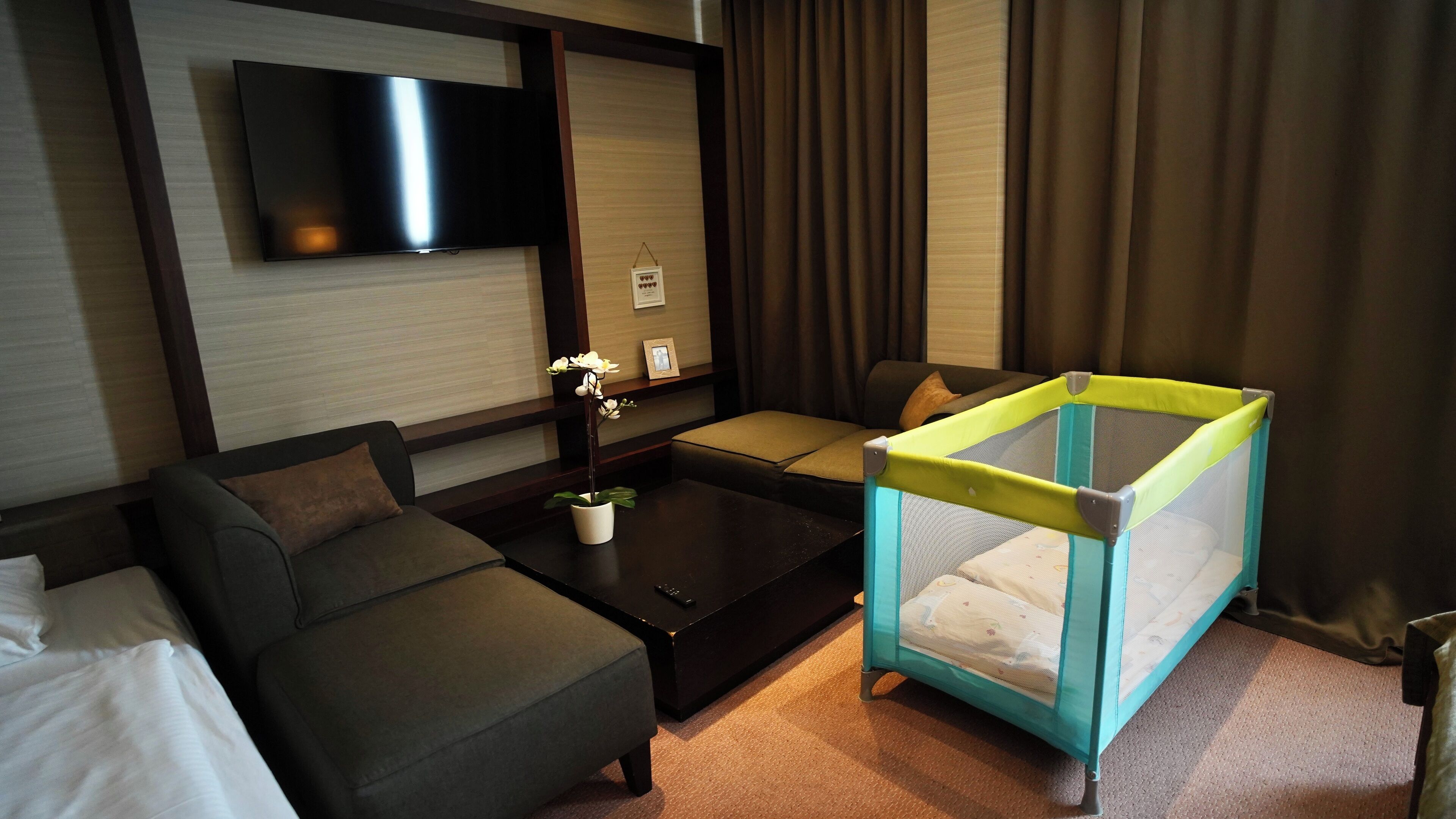 Habitación de hotel con sofá, una mesa de centro, televisor en la pared, una mesita con una planta y un corralito para bebé.