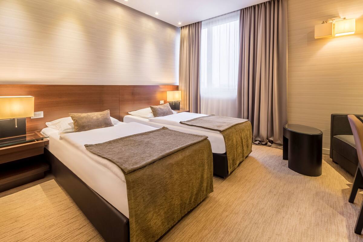 A hotel room with two beds, a nightstand, a lamp, a chair, and a window with curtains.