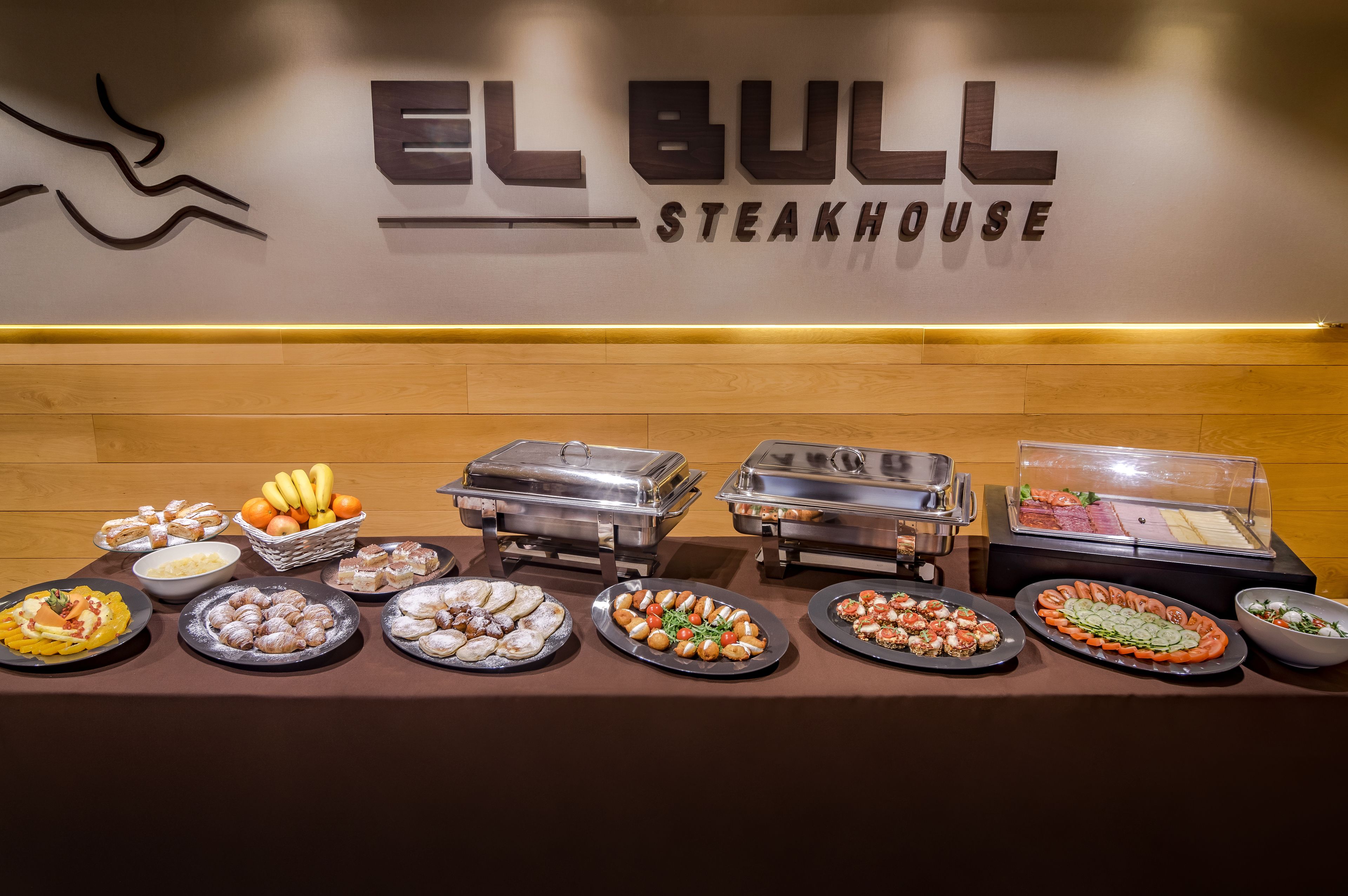 A buffet table with various food items and a wall with the sign "EL BULL STEAKHOUSE".