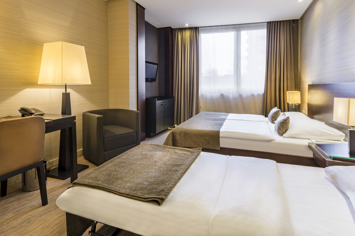 A hotel room with two beds, a desk with a telephone, a chair, a television, and a window with curtains.