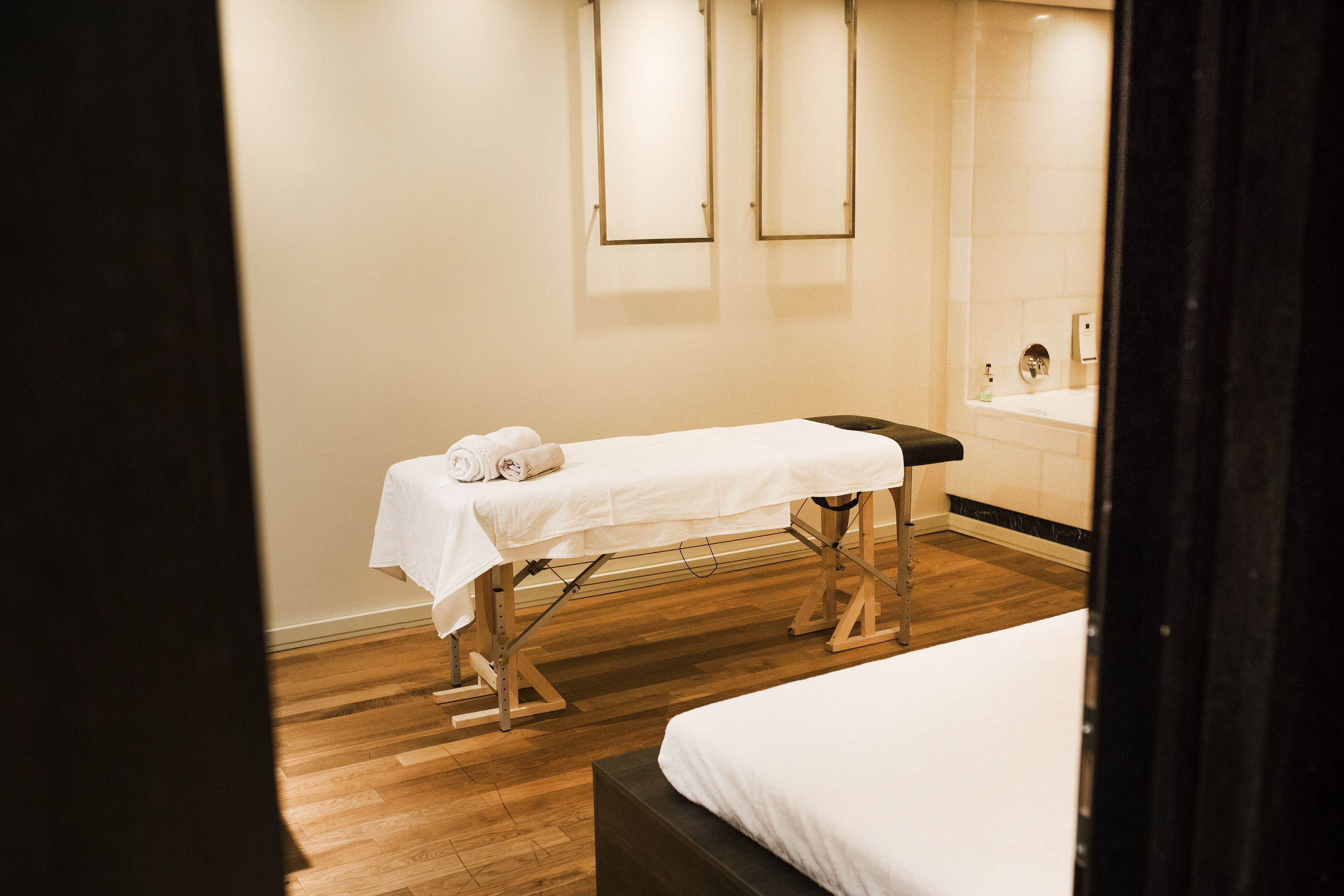A clean, modern room with a wooden floor, a bed, a massage table with towels, and a large mirror.