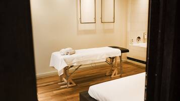A clean, modern room with a wooden floor, a bed, a massage table with towels, and a large mirror.