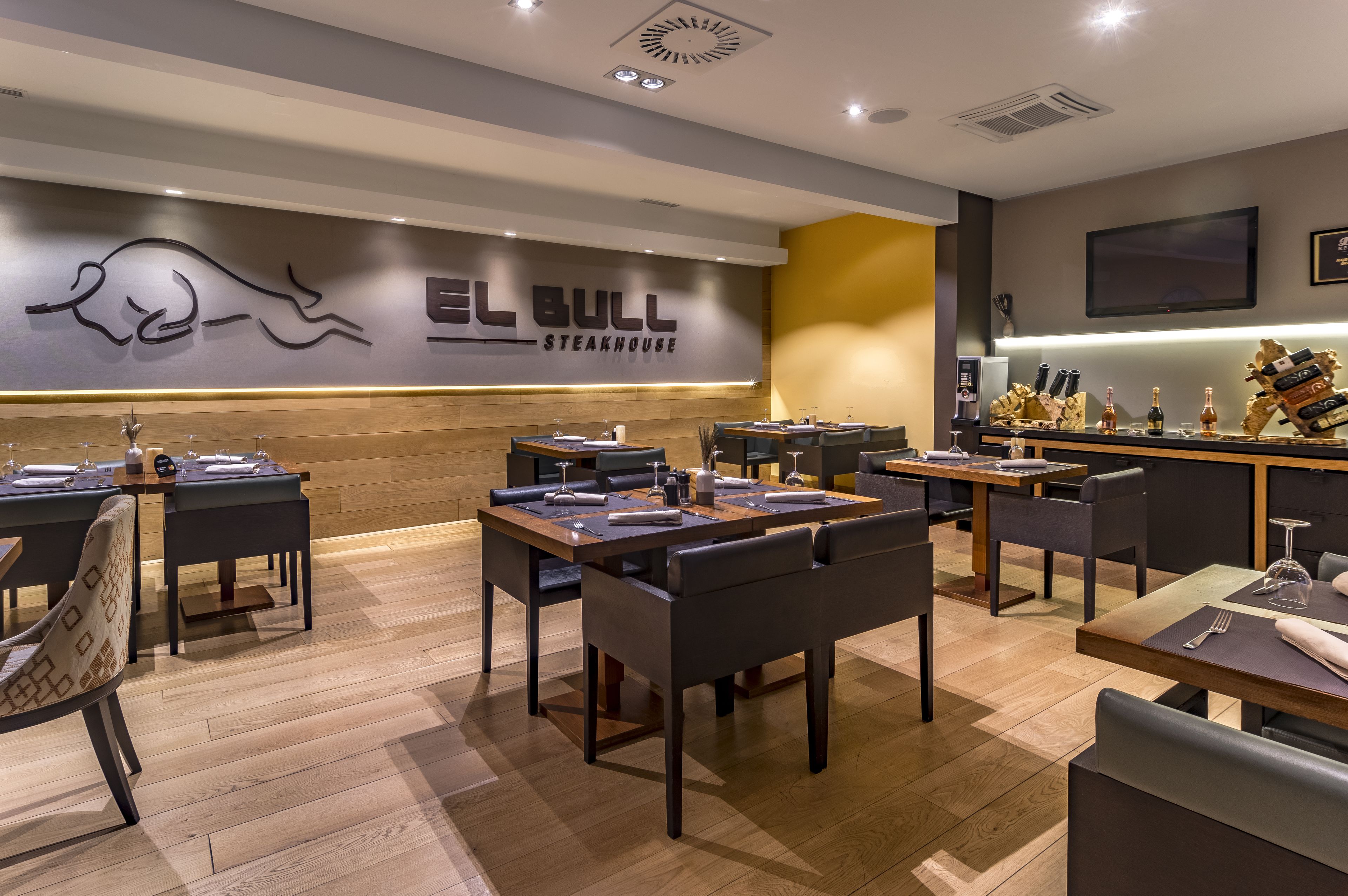 L’intérieur d’un restaurant, avec de nombreuses tables et chaises, un téléviseur fixé au mur et un bar où sont exposées des bouteilles.