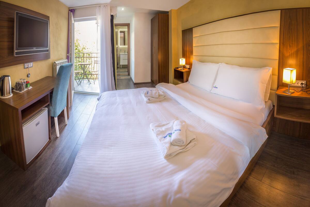 A hotel room with a large bed, a TV mounted on the wall, a desk with a kettle, and a small refrigerator.