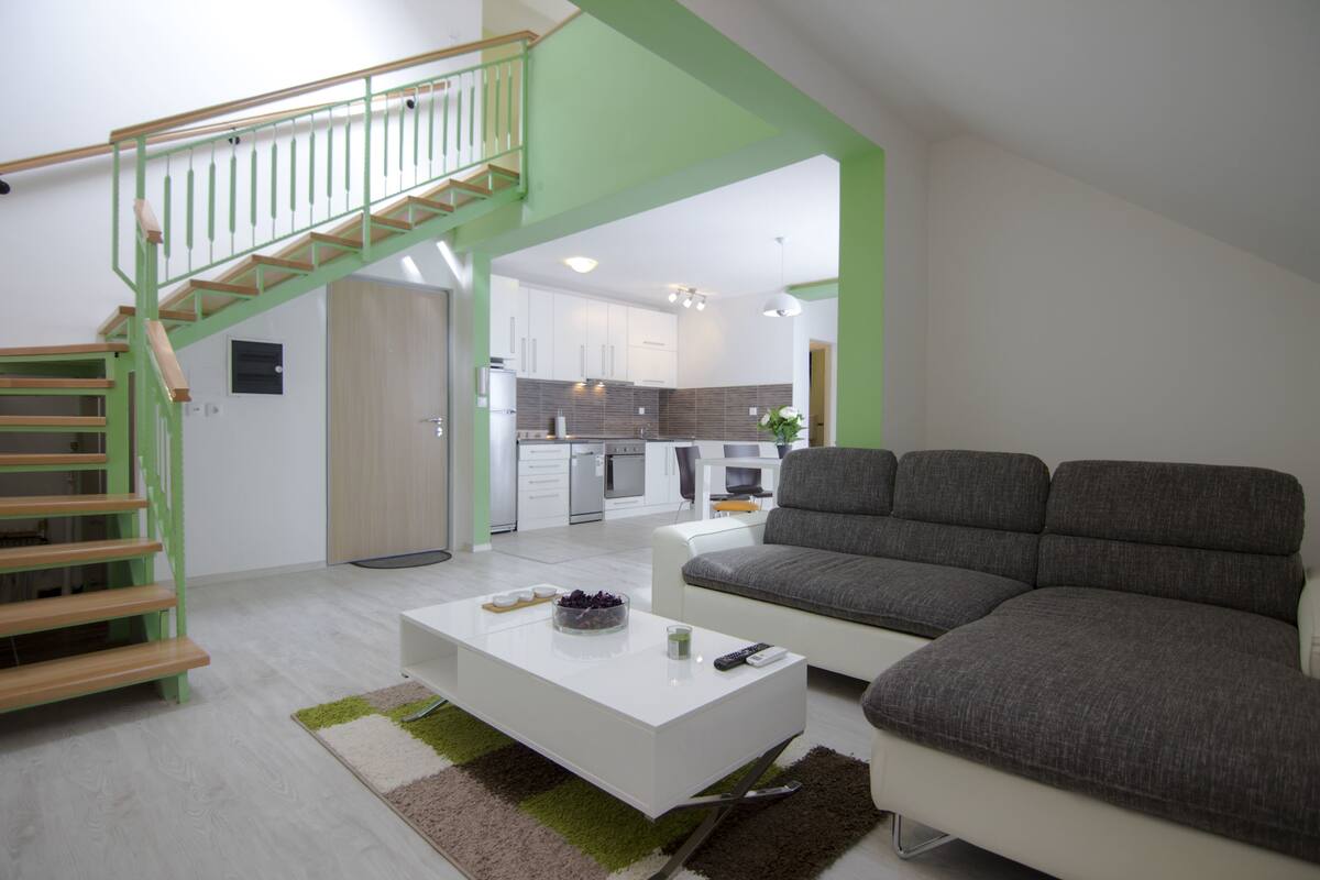 A modern living room with a staircase, a grey sofa, a white coffee table, and a kitchen in the background.