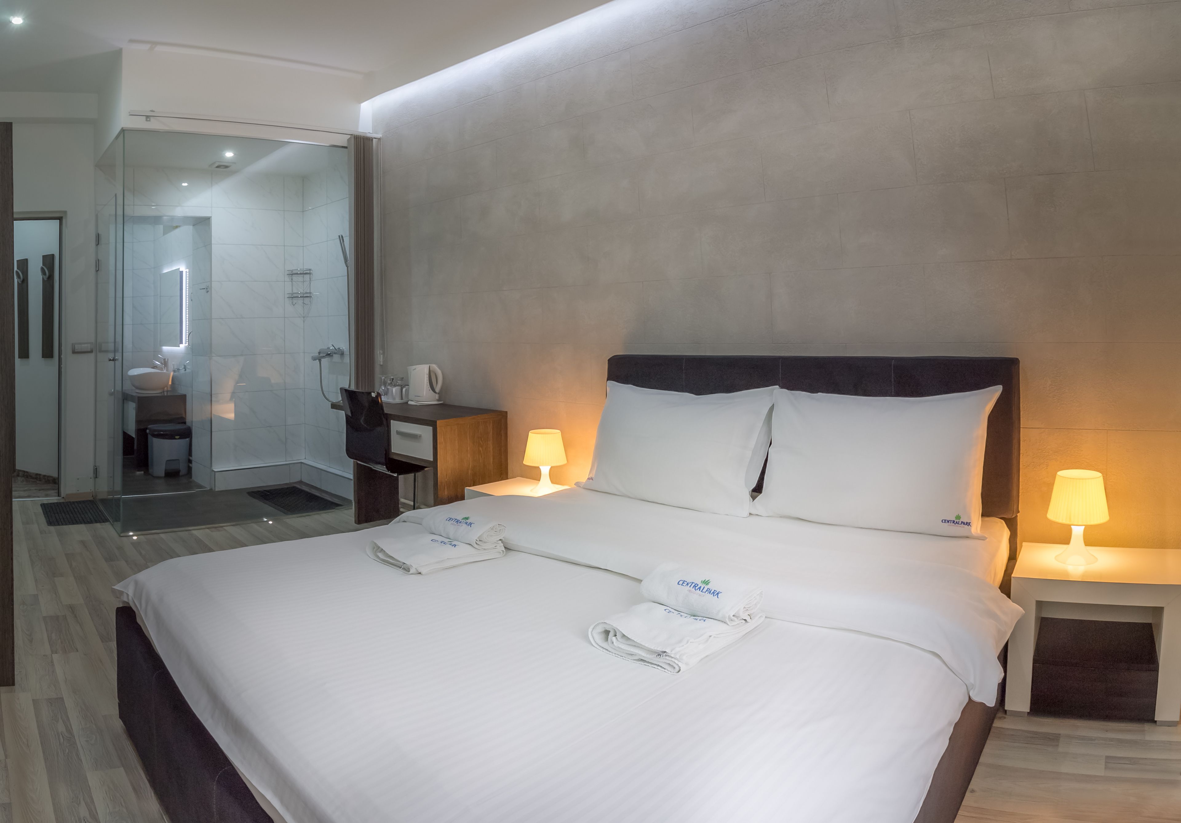 Habitación de hotel con una cama grande, dos lámparas de noche, cabina de ducha de vidrio, suelo de madera y una mesita con lámpara.