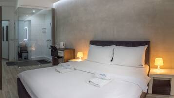Habitación de hotel con una cama grande, dos lámparas de noche, cabina de ducha de vidrio, suelo de madera y una mesita con lámpara.