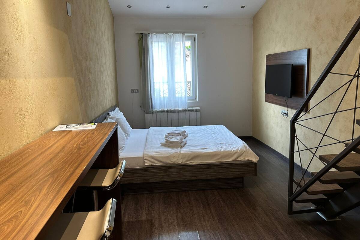 A compact bedroom with a bed, a wooden desk, a TV mounted on the wall, and a staircase.