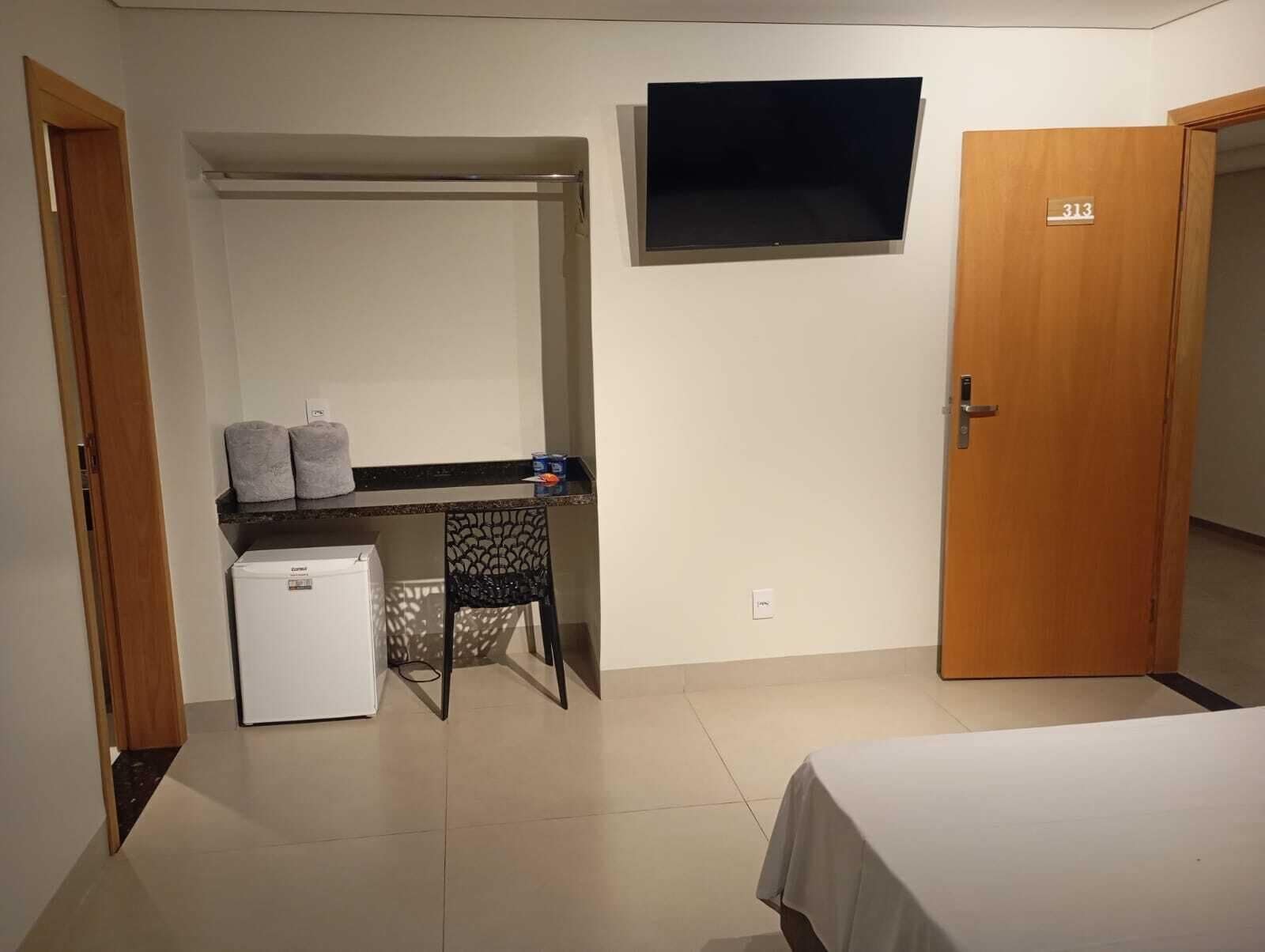 Una habitación compacta con cama, escritorio con silla, refrigerador pequeño, TV montada en la pared y una puerta con número.
