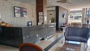 A hotel reception area with a marble counter, seating area with chairs and a table, and a television mounted on the wall. - Bravo City Hotel Dourados (Dourados)