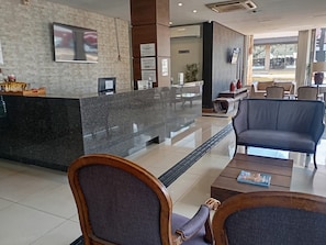 A hotel reception area with a marble counter, seating area with chairs and a table, and a television mounted on the wall. - Bravo City Hotel Dourados (Dourados)
