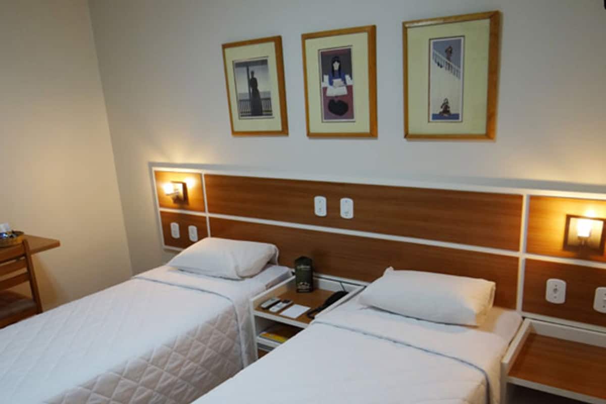 A hotel room with two beds, each with a wooden headboard and bedside lamps. There are framed pictures on the wall above the beds.