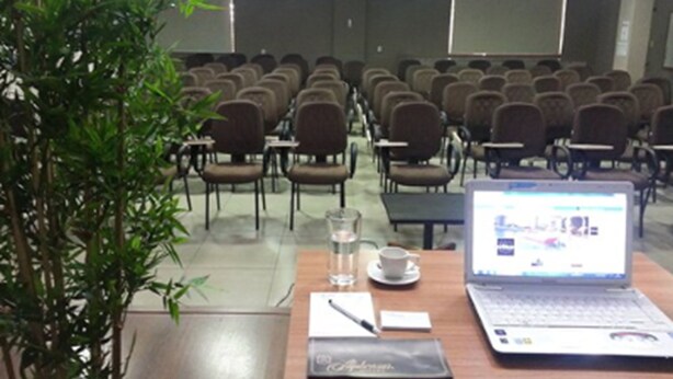 A conference room with rows of chairs, a laptop, a glass of water, and a notebook on a table. - Bravo City Hotel Dourados (Dourados)