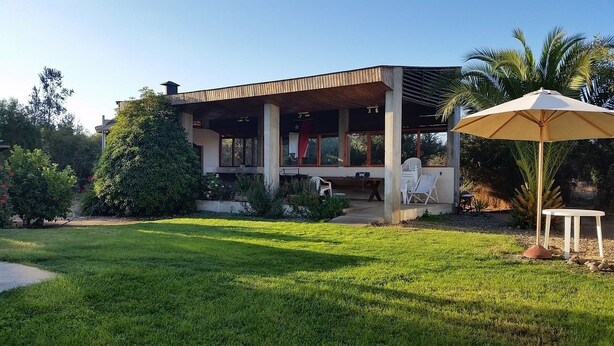Ett modernt hus med en inglasad veranda, en gräsmatta och en uteplats med stolar och ett parasoll.