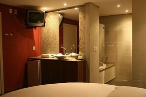 A hotel bathroom with a large mirror, a sink, a shower, and a television mounted on the wall. - Bangalô Motel - Adults Only (Santa Maria)