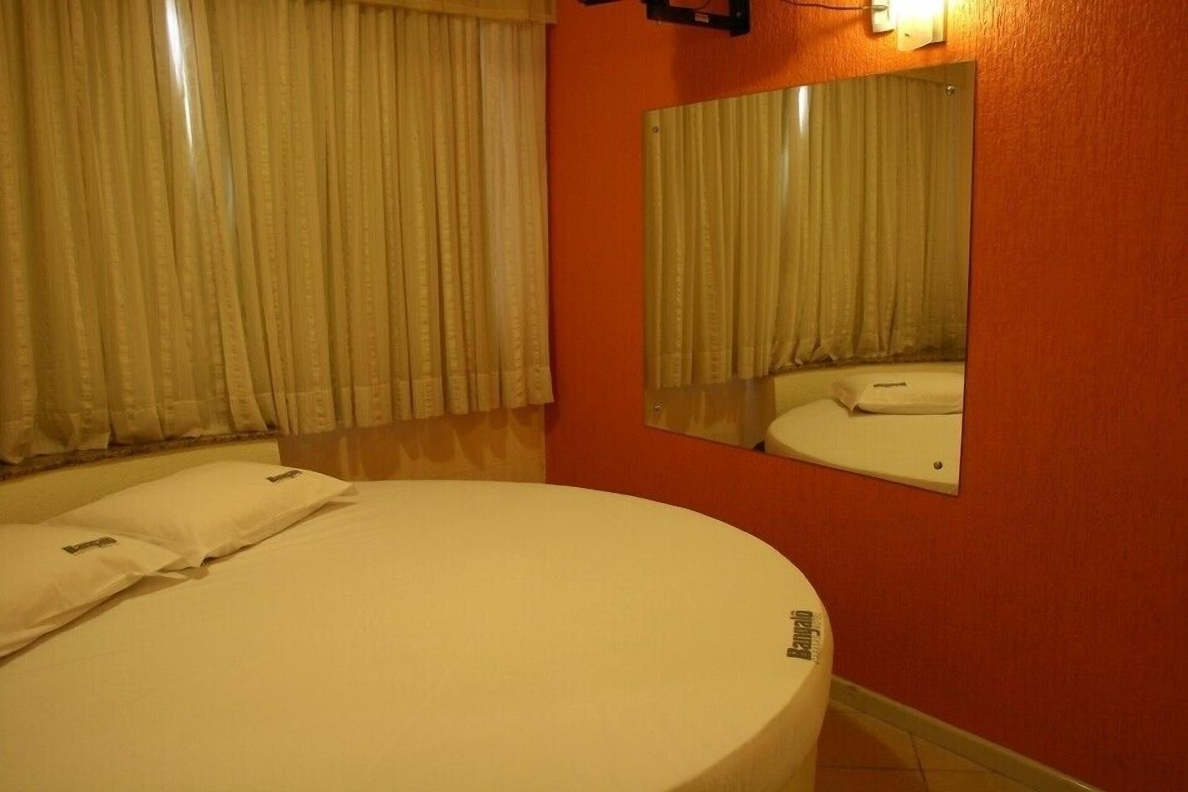 Quarto de hotel com uma cama grande, uma mesa de cabeceira redonda, um espelho e uma janela com cortinas.