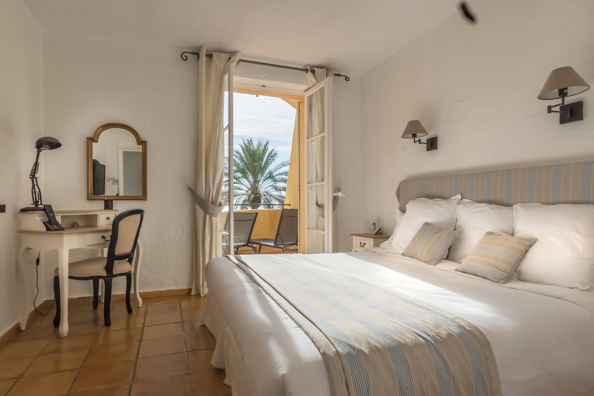 Ein Schlafzimmer mit Bett, Schreibtisch, Stuhl, Spiegel und Blick auf Palmen durch ein offenes Fenster.