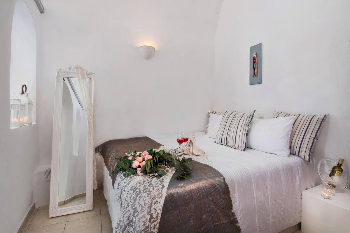 Un dormitorio con una cama, una mesita de noche con una botella y una vela, un espejo, y una lámpara de pared.