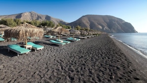 Una playa con hileras de tumbonas y sombrillas de paja, cielo despejado y montañas al fondo.