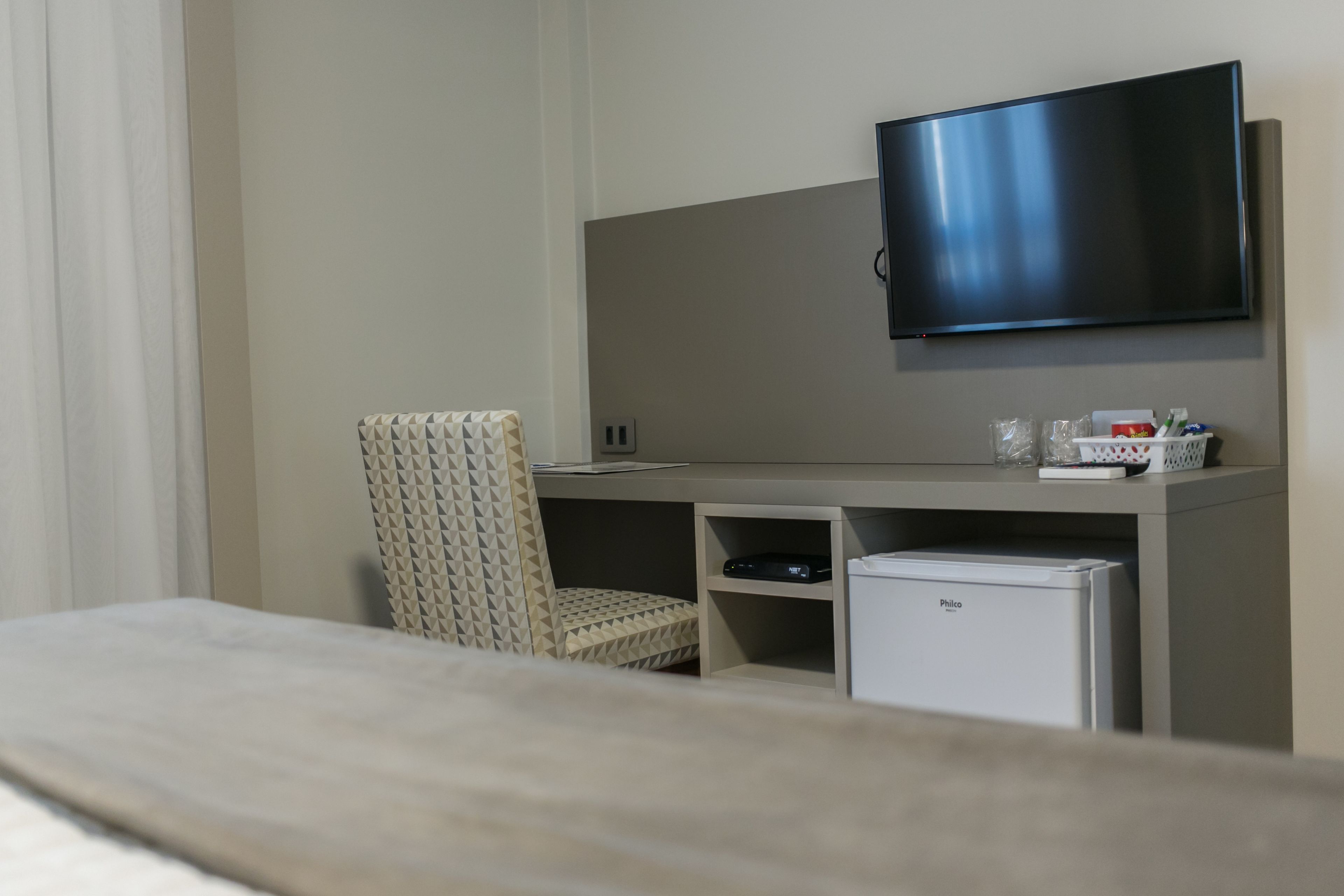 Habitación de hotel con cama, una silla, escritorio con televisor de pantalla plana, refrigerador pequeño y estante con artículos de aseo.