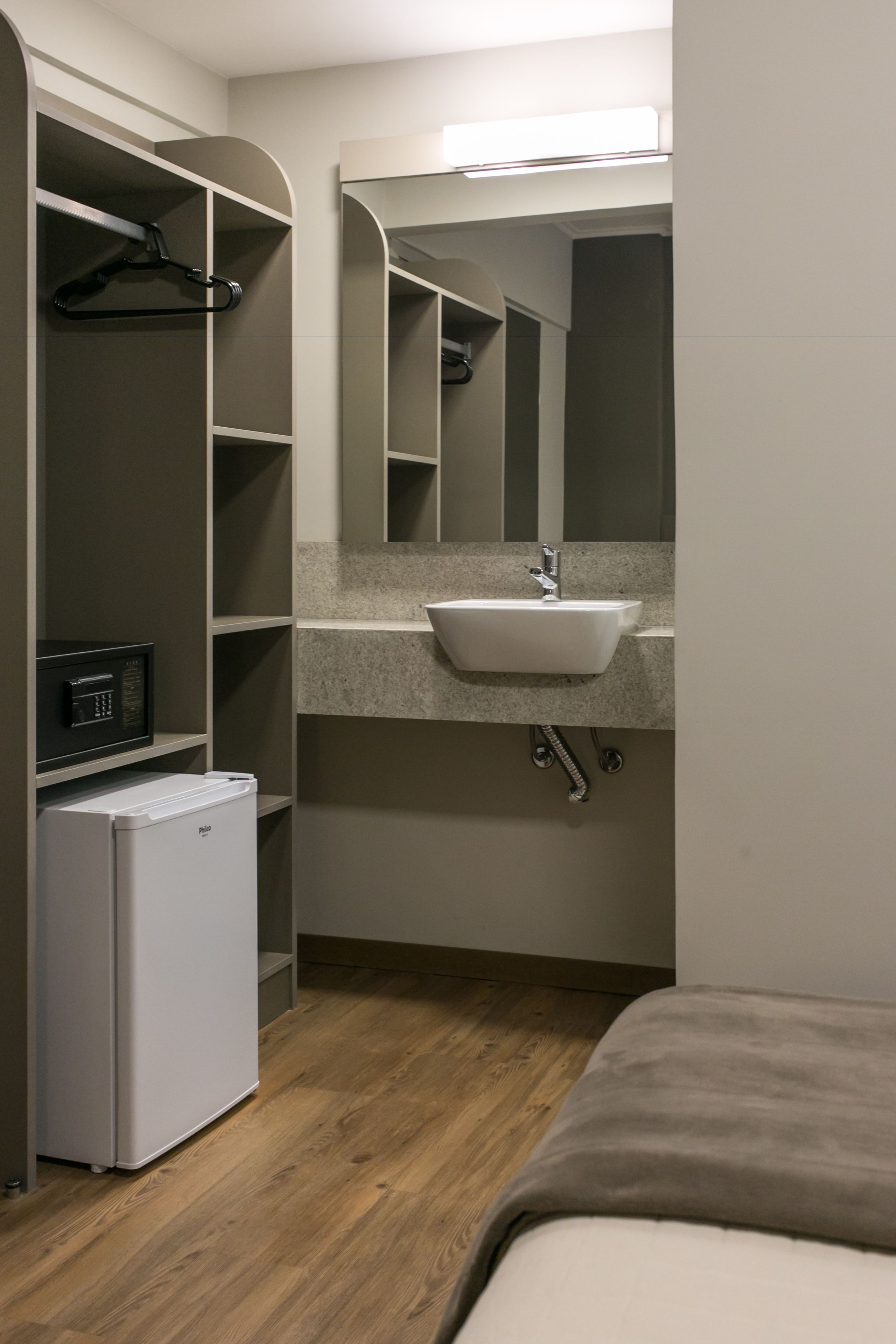 Ein kleines Hotelzimmer mit integriertem Kleiderschrank, einem kleinen Kühlschrank, einem Waschbecken und einem Spiegel.