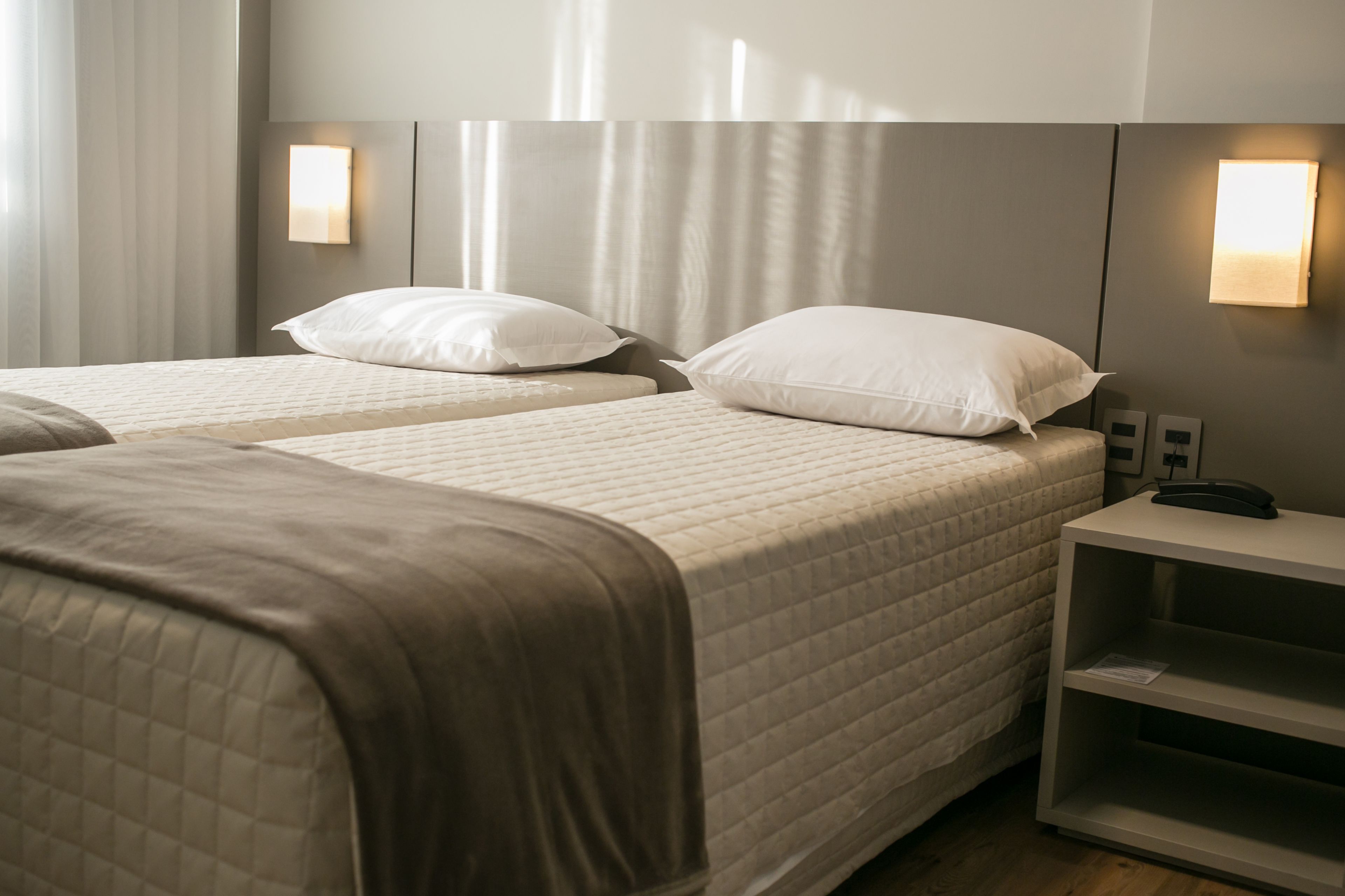 Une chambre d’hôtel avec deux lits, chacun recouvert d’une courtepointe blanche matelassée et agrémenté d’oreillers, une table de chevet avec une lampe et un luminaire fixé au mur.