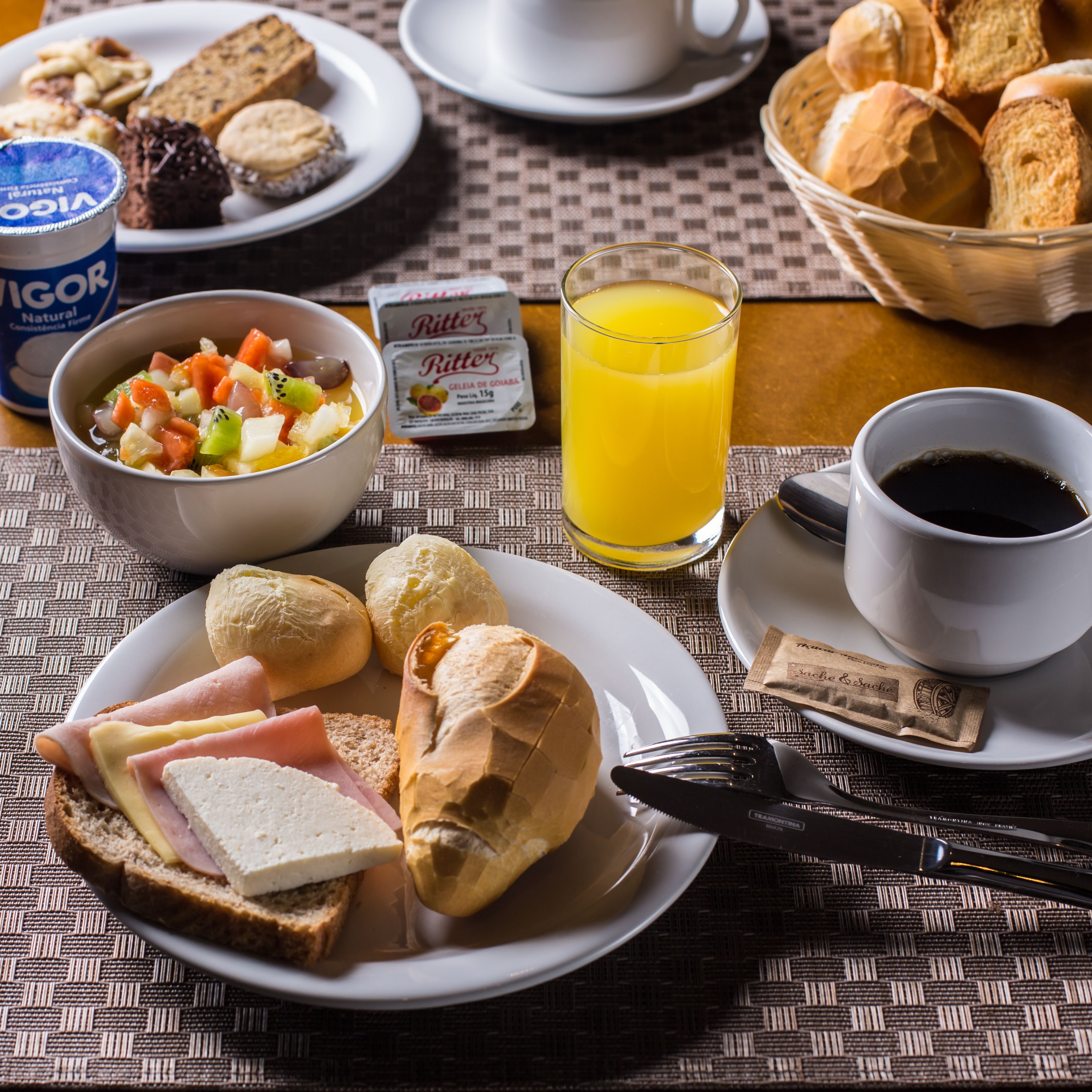 A breakfast setting with a plate of ham and cheese, croissants, a bowl of fruit salad, a glass of orange juice, and a cup of coffee.