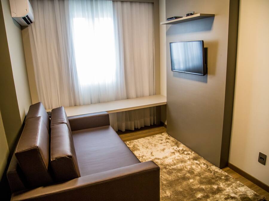 Una sala moderna con un sofá de cuero marrón, un televisor de pantalla plana montado, una ventana con cortinas translúcidas y un banco empotrado.