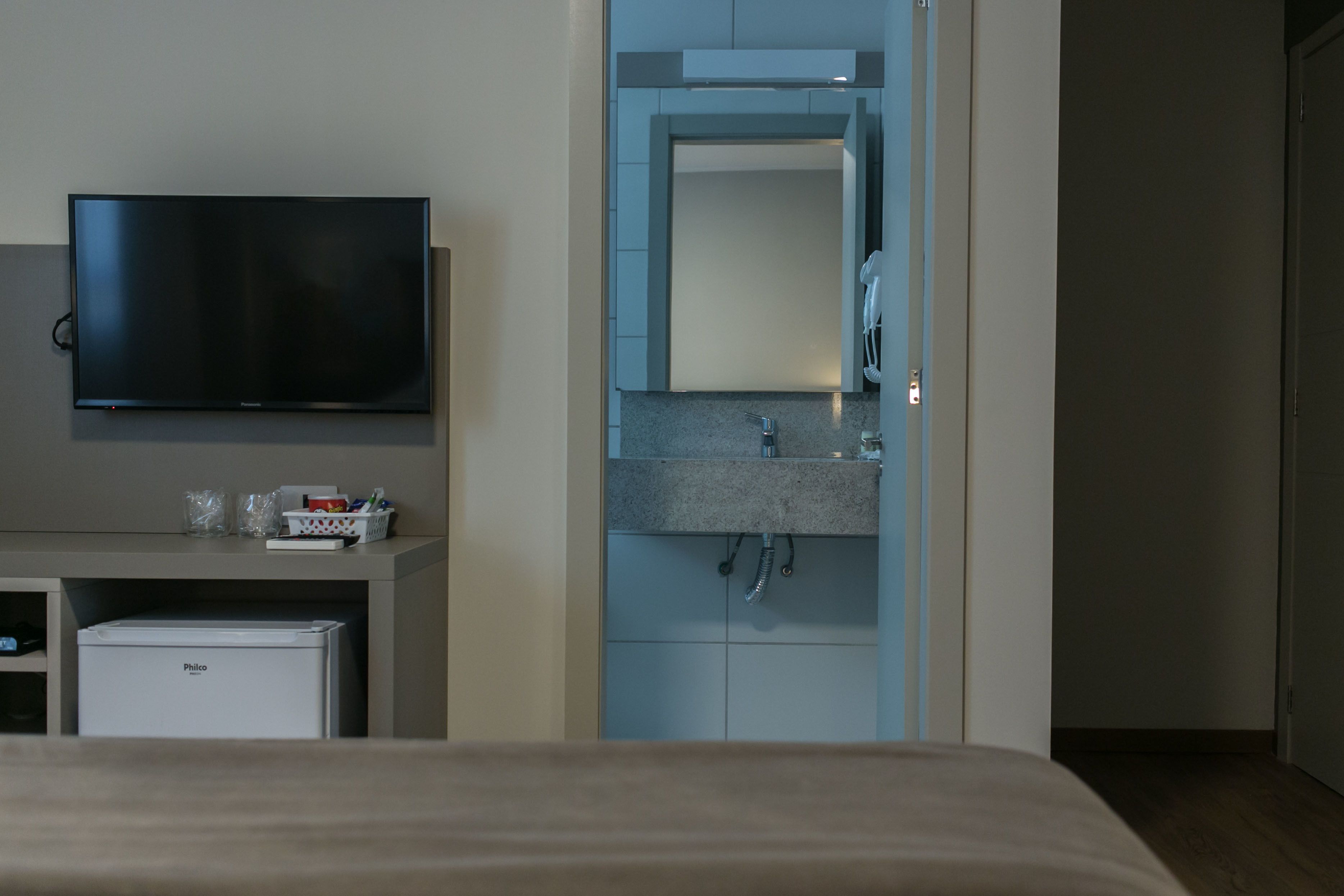 Quarto de hotel com televisão de tela plana, minibar, banheiro com pia e espelho, e uma porta à direita.