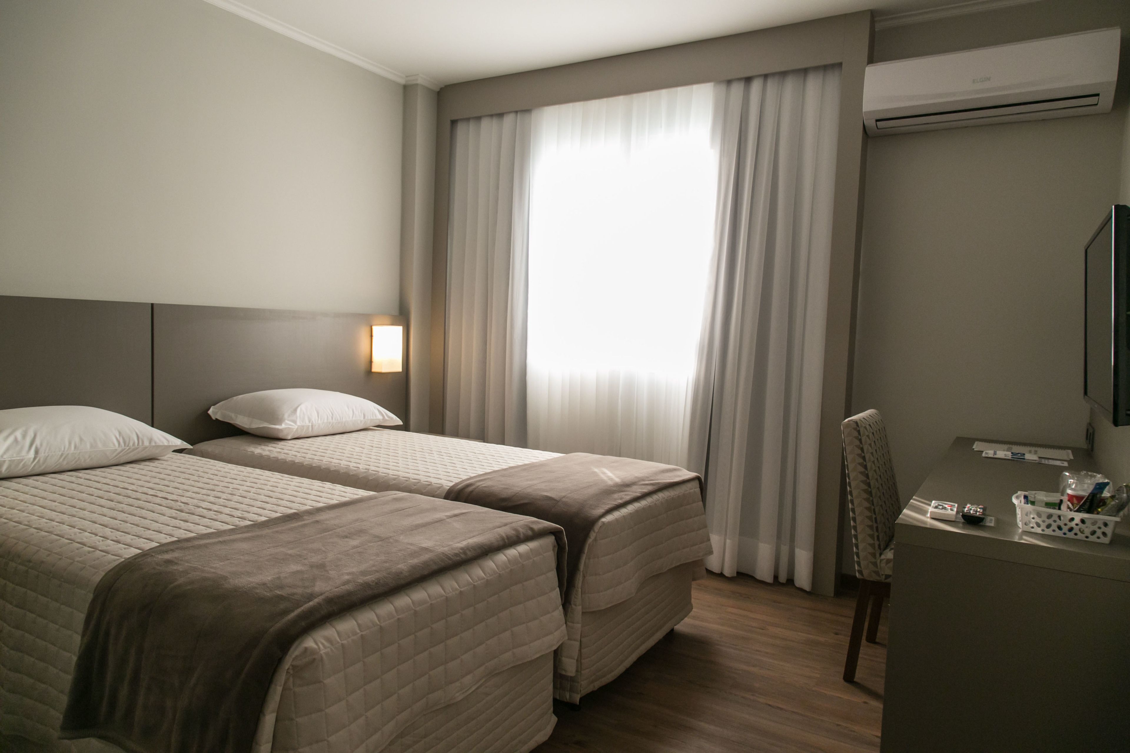 Habitación de hotel con dos camas, un escritorio, una silla y un ventanal grande con cortinas.