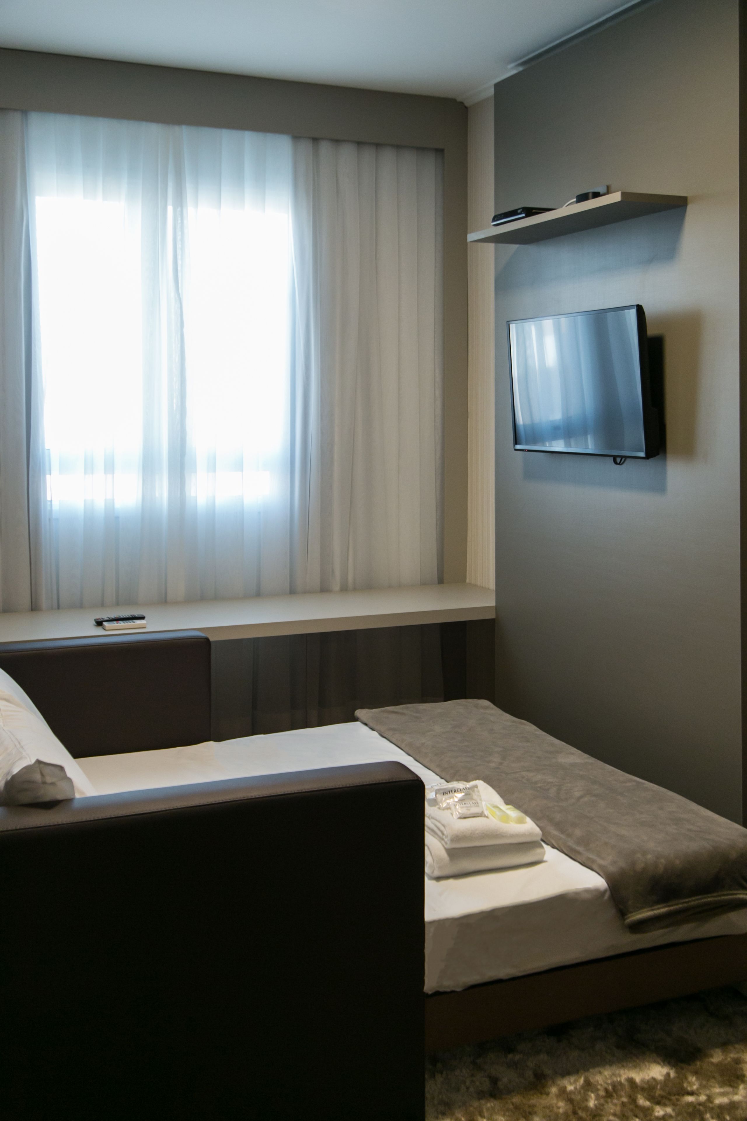 A hotel room with a bed, a TV mounted on the wall, a window with curtains, and a small shelf above the bed.