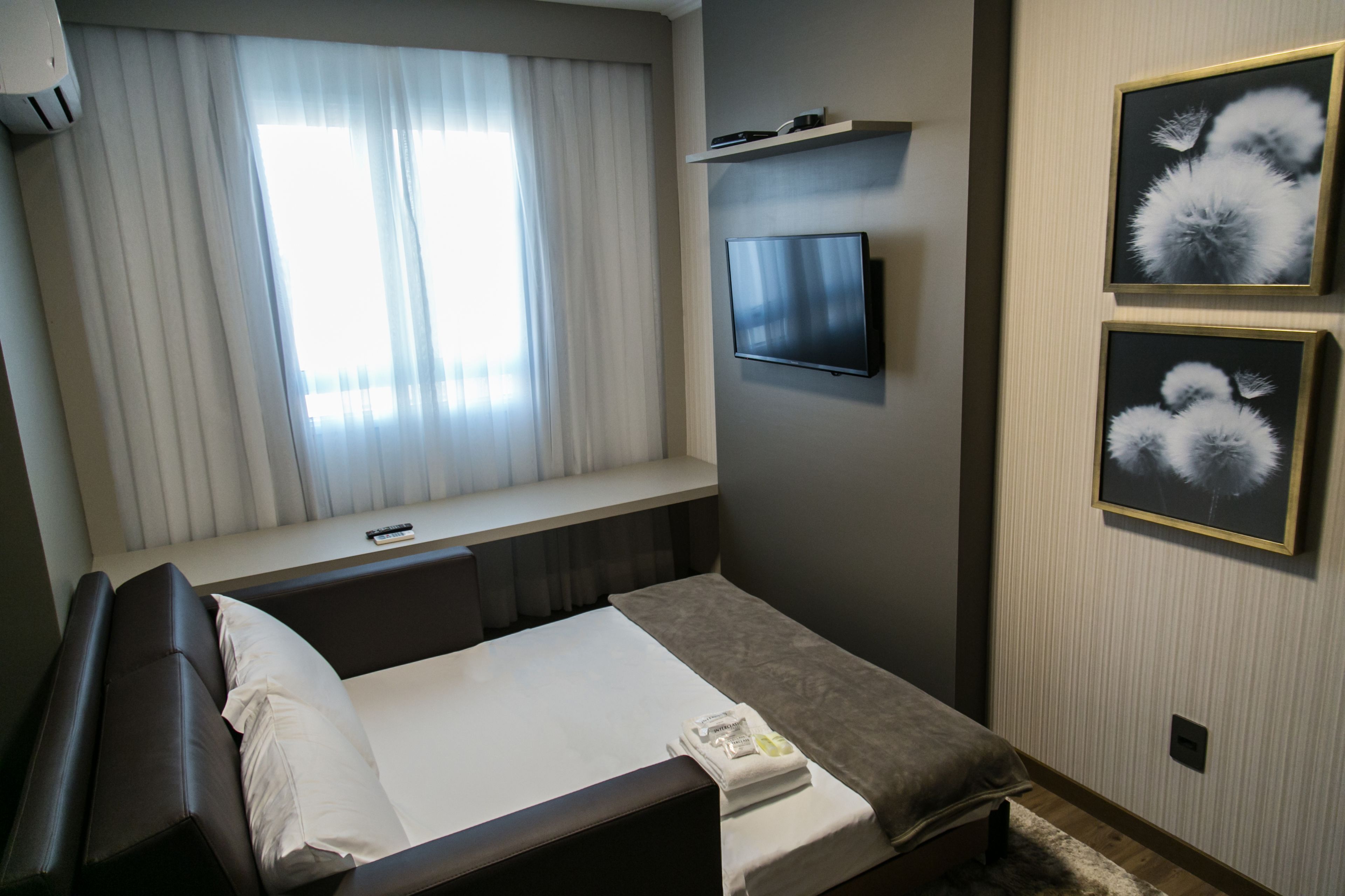 Una habitación de hotel con cama, televisor montado en la pared y dos cuadros enmarcados.