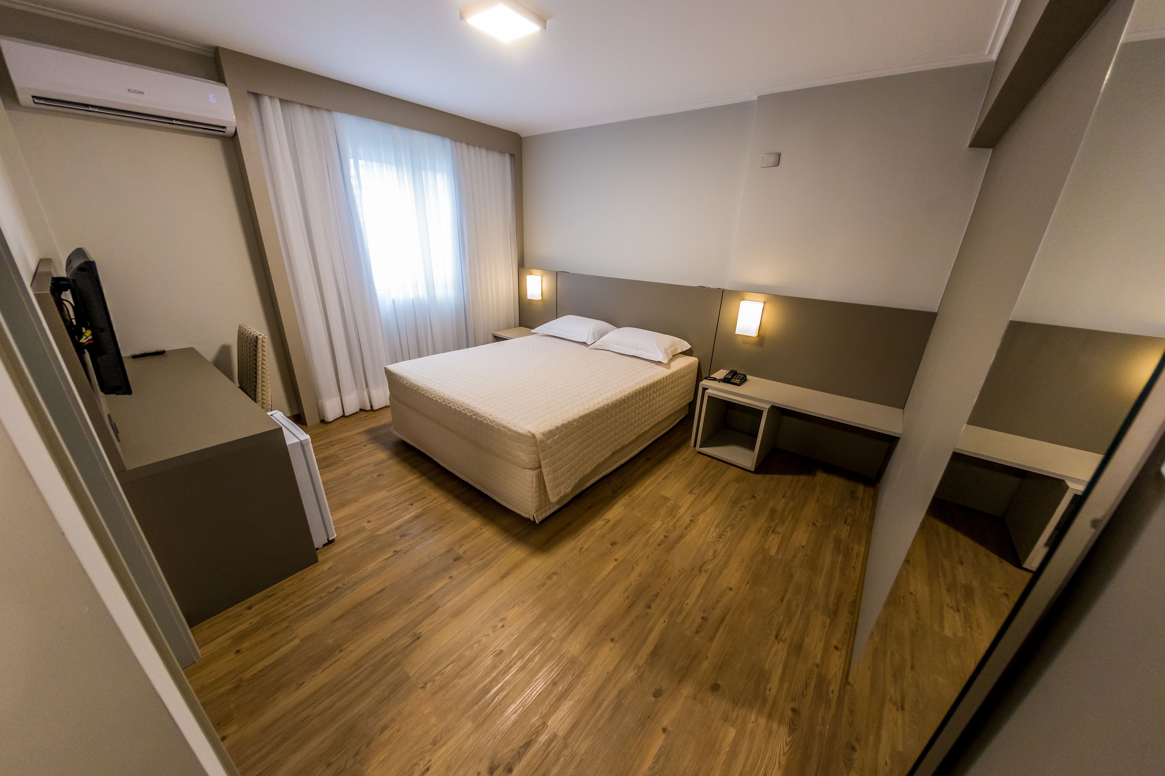 A hotel room with a bed, bedside tables, a desk, and an air conditioning unit.