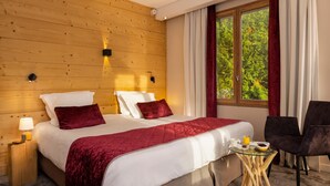 A hotel room with a wooden wall, a large bed with white and red bedding, a small table with breakfast items, and a window with a view of greenery. - Hôtel Les Grillons (Talloires-Montmin)