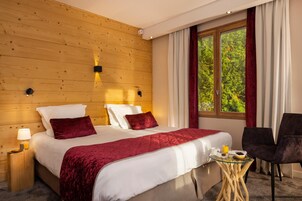 Quarto de hotel com parede de madeira, uma cama grande com roupa de cama branca e vermelha, uma mesa pequena com itens de café da manhã e uma janela com vista para o verde.