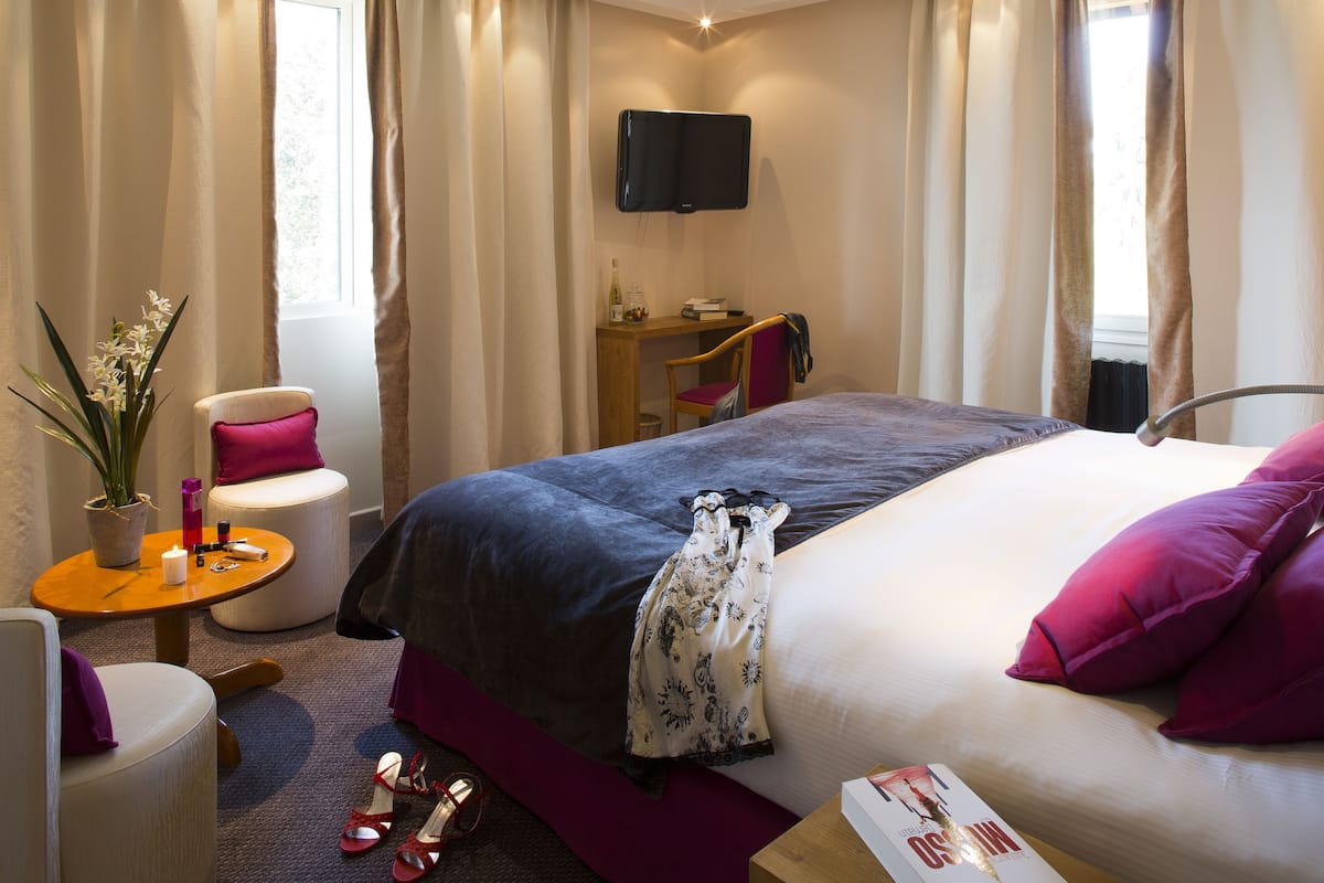A hotel room with a bed, a desk, a chair, a small table, and a potted plant.