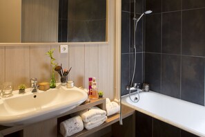 A bathroom with a white sink, a mirror, a shower with a handheld showerhead, and a shelf with towels and toiletries. - Hôtel Les Grillons (Talloires-Montmin)