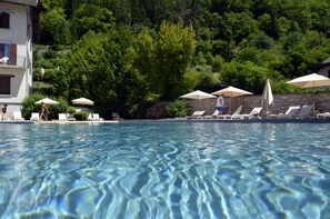 A swimming pool with clear water, surrounded by lounge chairs and umbrellas, with a forested hillside in the background. - Hôtel Les Grillons (Talloires-Montmin)