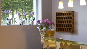 A reception area with a glass counter, potted plants, and a wooden shelf with bottles. - Hôtel Les Grillons (Talloires-Montmin)