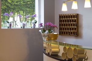 Uma área de recepção com balcão de vidro, plantas em vasos e prateleira de madeira com garrafas.