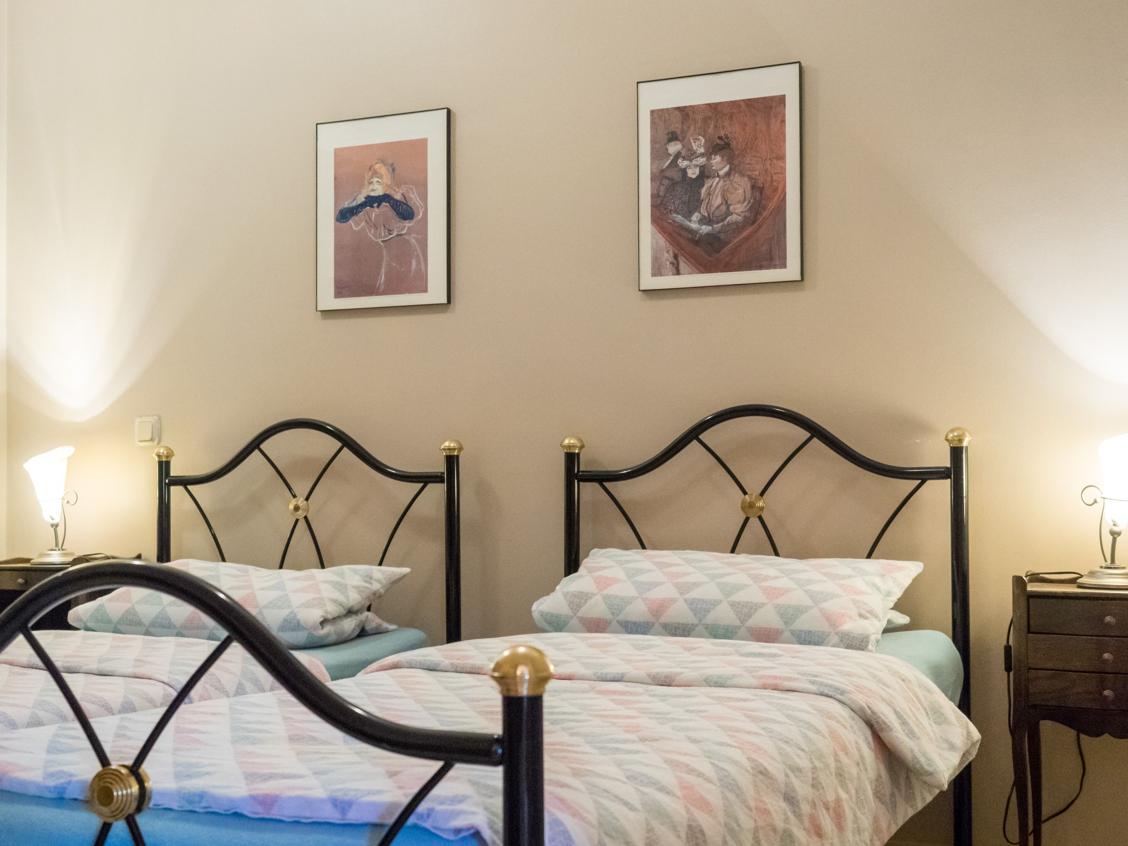 Ein Schlafzimmer mit zwei Betten, jeweils mit schwarzem Metallgestell und goldenen Akzenten, sowie einer gemusterten Bettdecke. Es gibt zwei Nachttische mit Lampen, und die Wände sind mit gerahmten Kunstwerken geschmückt.