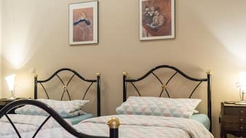 Ein Schlafzimmer mit zwei Betten, jeweils mit schwarzem Metallgestell und goldenen Akzenten, sowie einer gemusterten Bettdecke. Es gibt zwei Nachttische mit Lampen, und die Wände sind mit gerahmten Kunstwerken geschmückt.