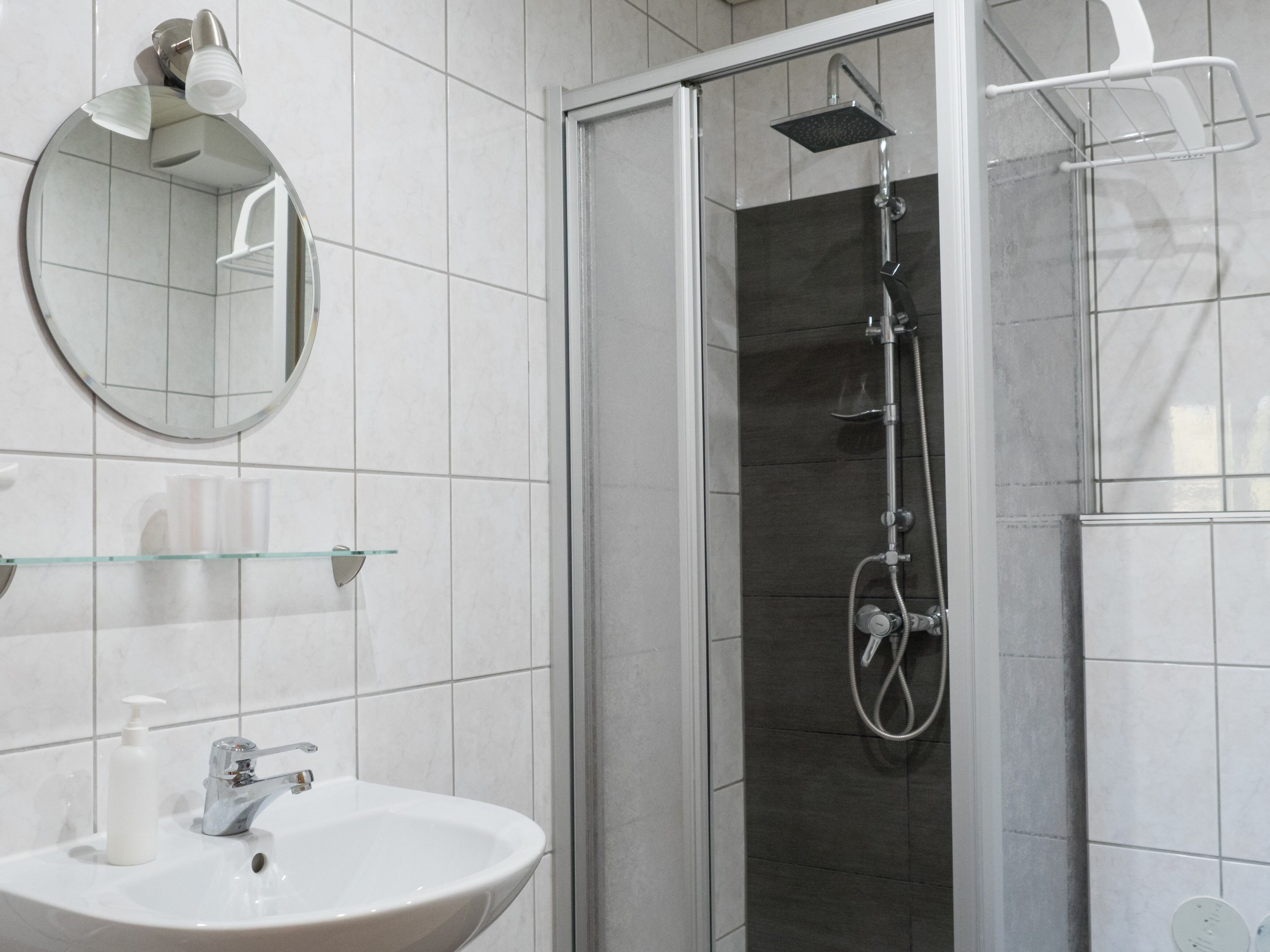 Un baño moderno con ducha envuelta en vidrio, un espejo redondo y un lavamanos blanco con dispensador de jabón.