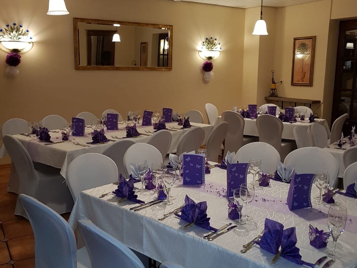 Un salón de banquetes con varias mesas redondas dispuestas para un evento formal, cada una con manteles blancos y centros de mesa morados.