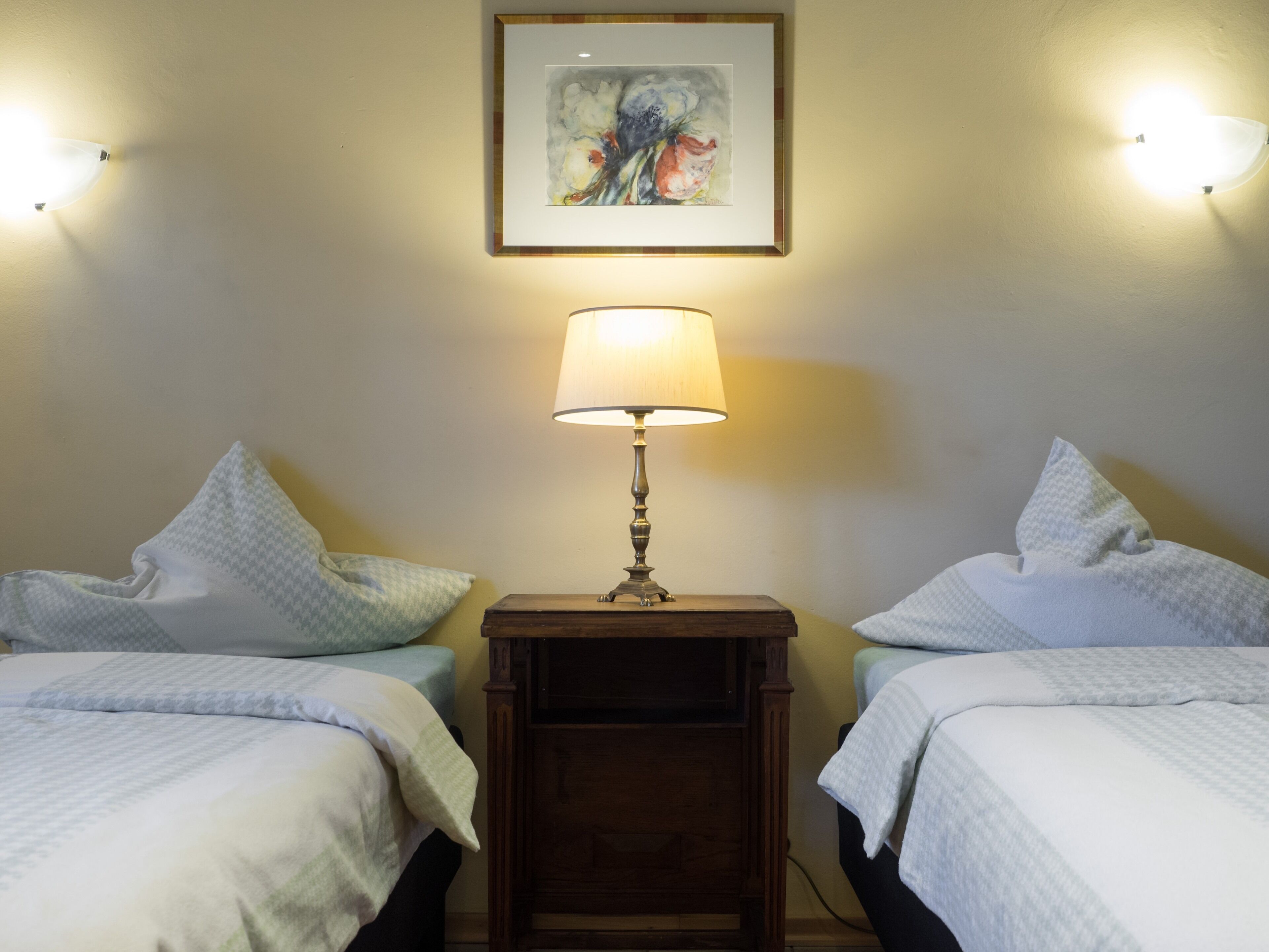 A hotel room with two beds, a bedside table with a lamp, and a framed artwork on the wall.