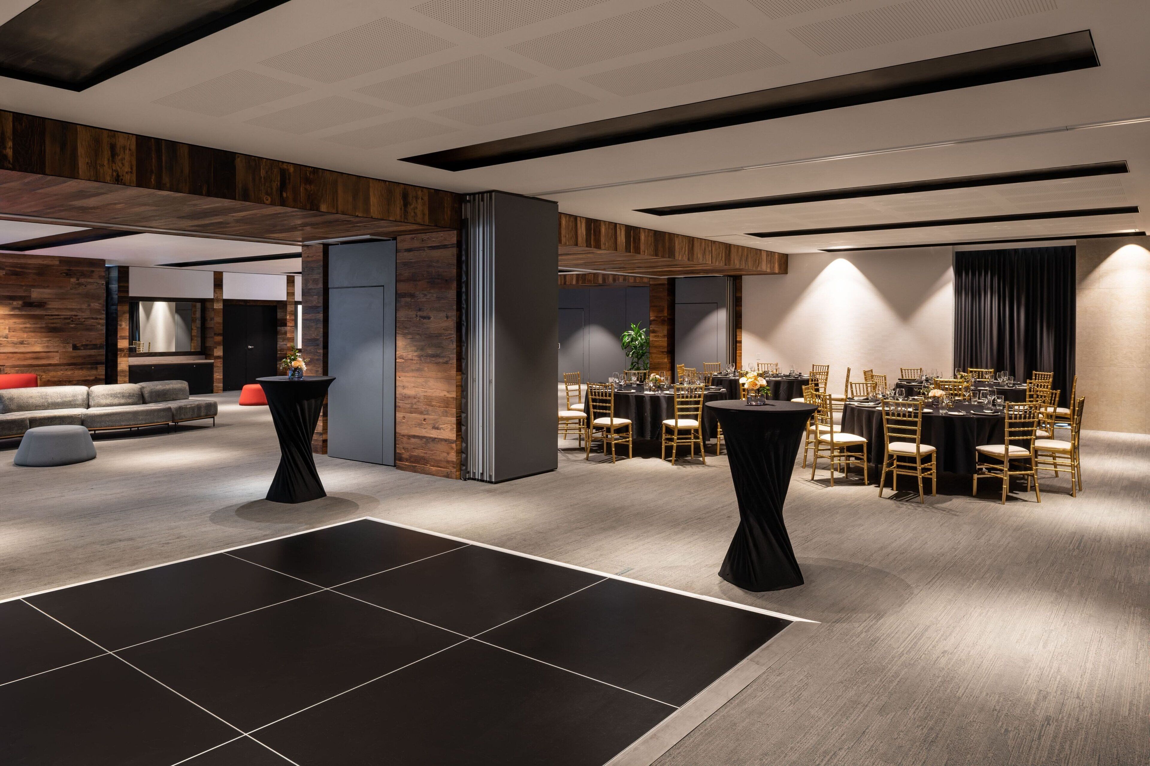 A modern hotel lobby with a mix of wooden and concrete elements, a central seating area, and a dining setup with round tables and chairs.