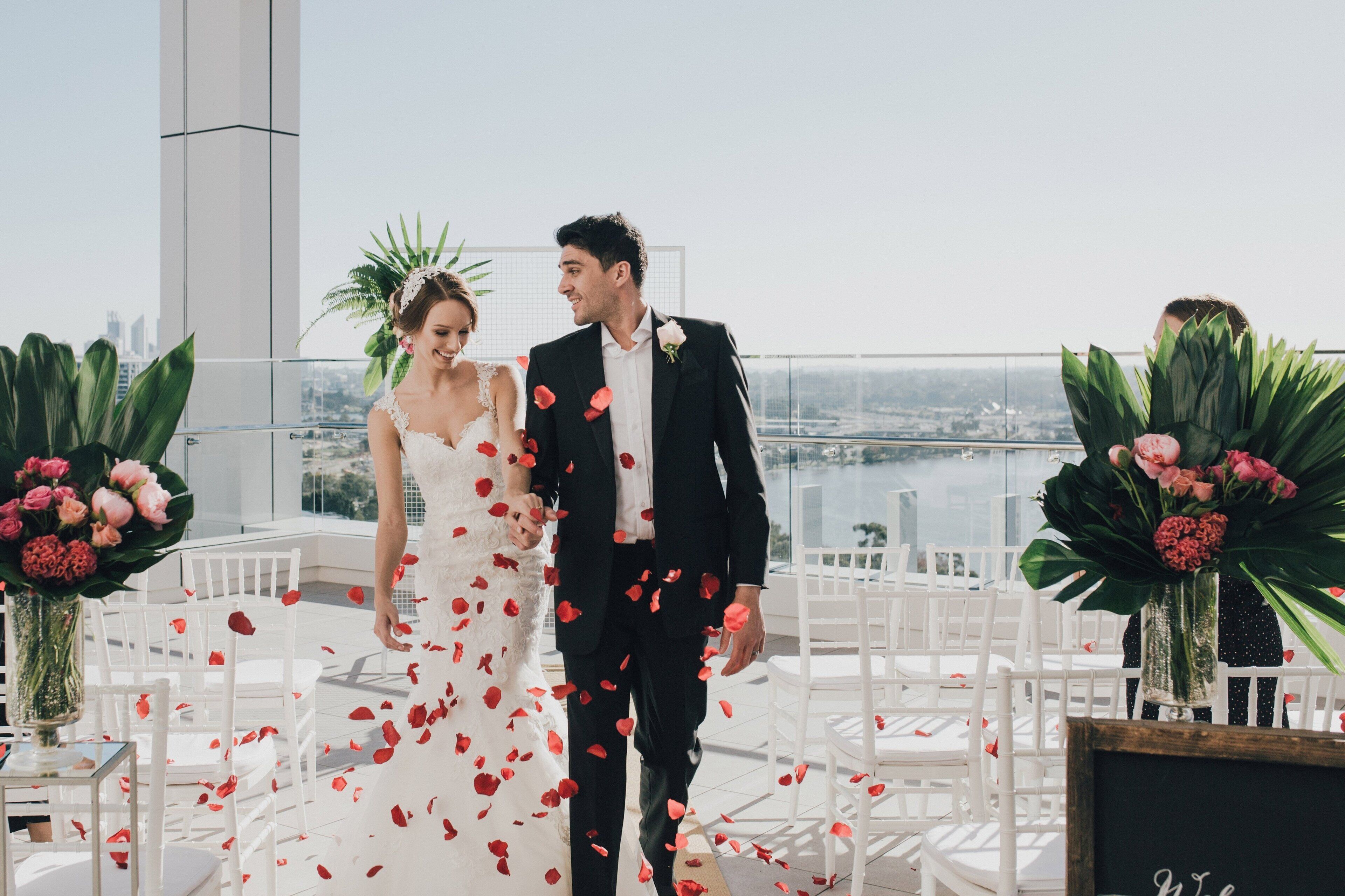 A couple in wedding attire on a rooftop terrace with a city view, white chairs, and floral arrangements.